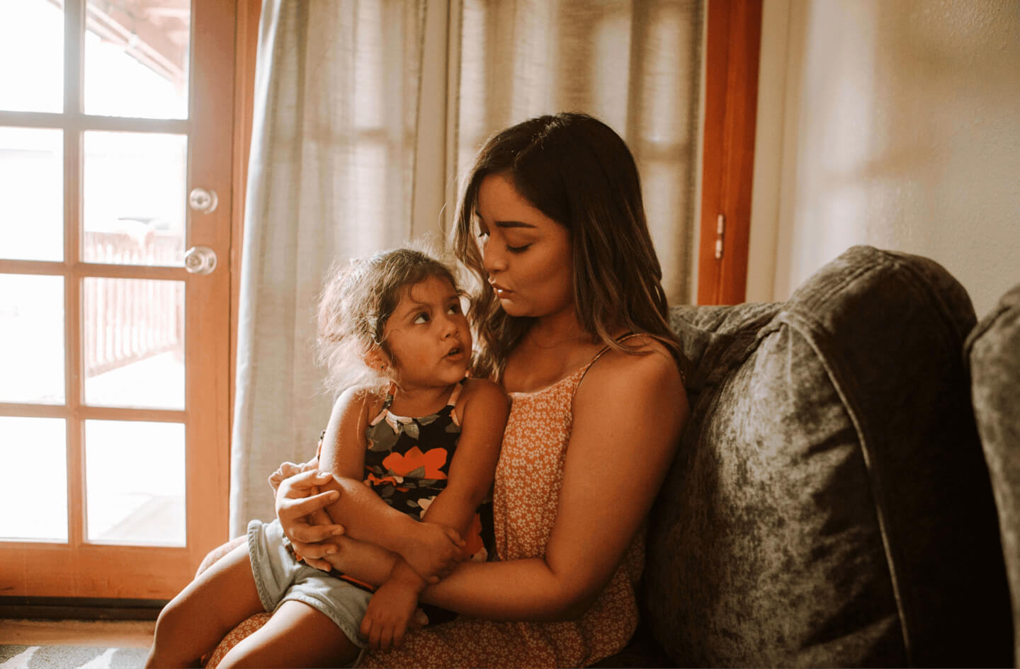 A mother holding her baby on a sofa beside a window