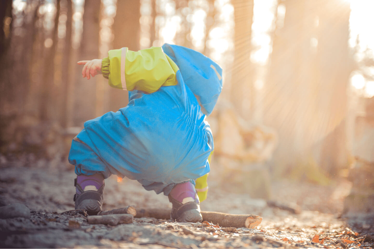 A toddler in a yellow raincoat crouching to explore the woodland floor