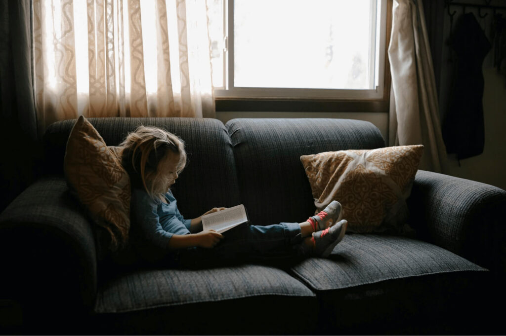 A young child sitting alone on a large sofa, looking at a book