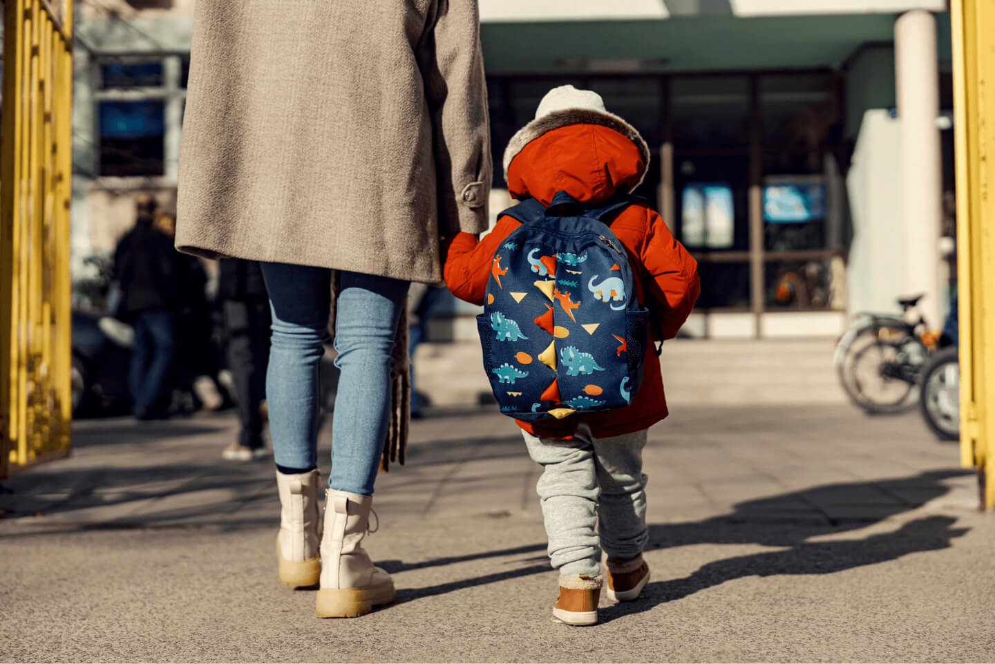 A young child in a red coat and backpack walking outdoors with an adult