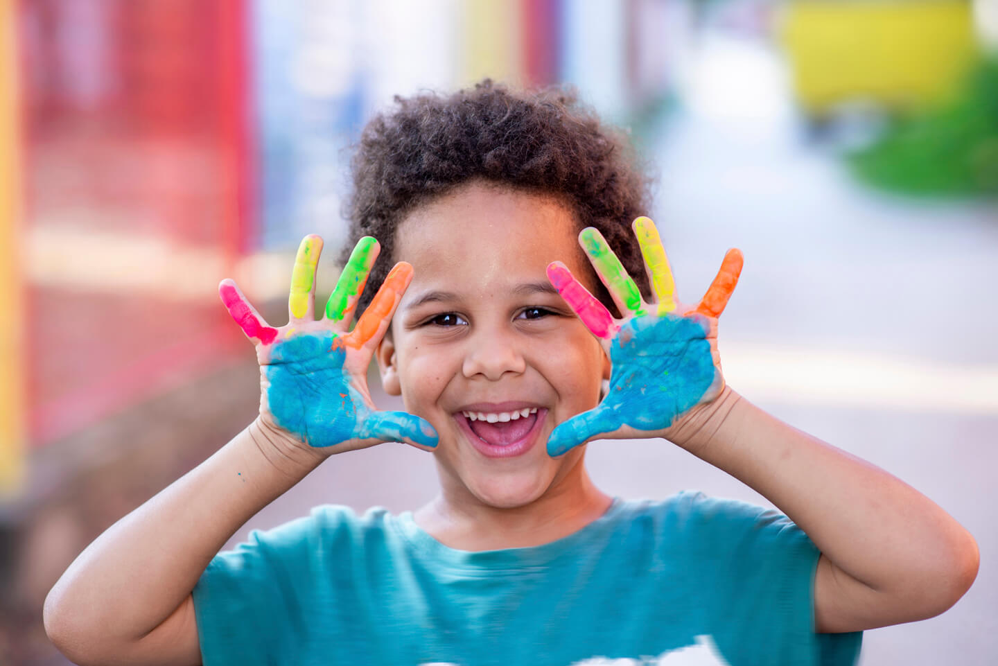 A young child laughing with brightly painted hands held up to the camera